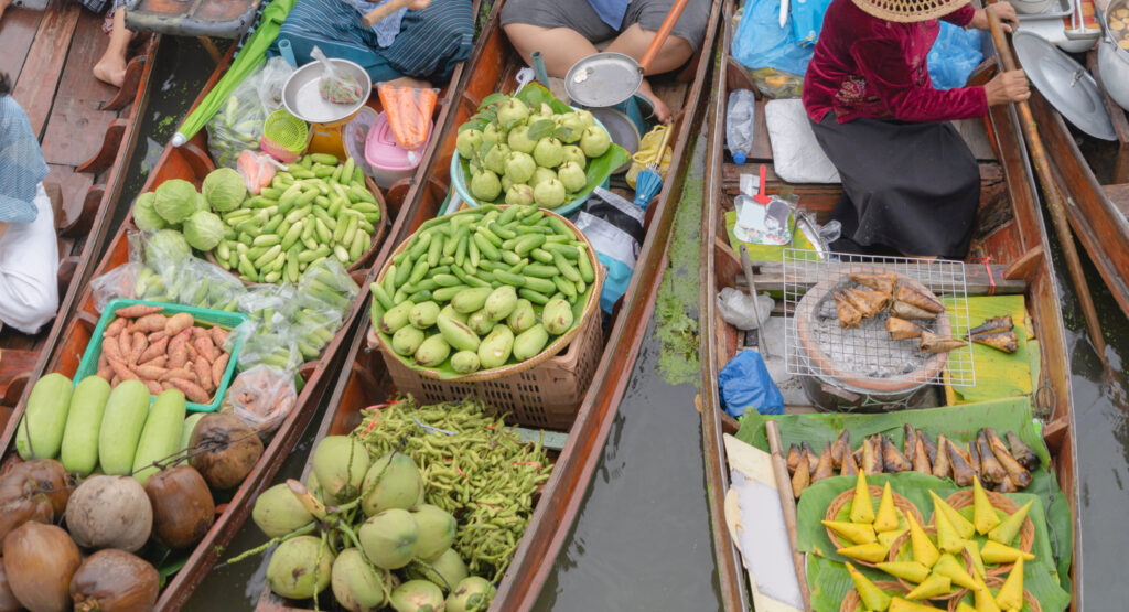 Übervolle Boote mit Gemüse, Kokosnüssen und gegrilltem Fisch am Damnoen Saduak Floating Market in Thailand.