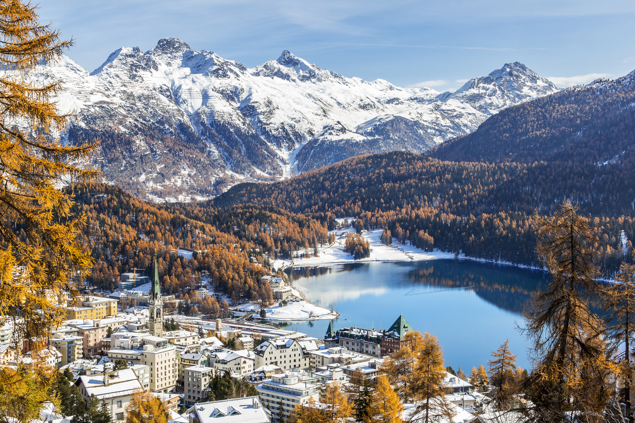 Blick über St. Moritz auf den stillen See, herbstliche Wälder und schneebedeckte Gipfel im Hintergrund.