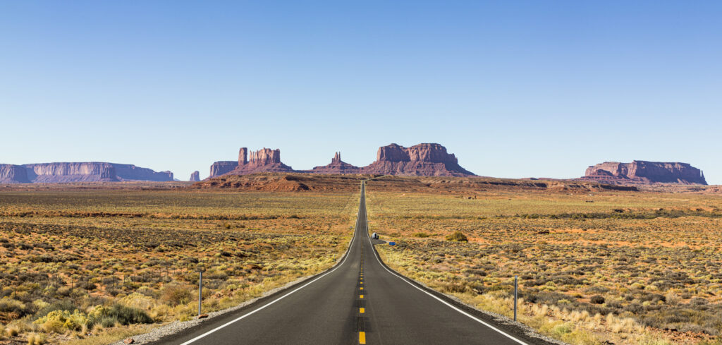Eine lange, gerade Straße führt durch die Weite von Monument Valley auf markante Tafelberge zu.