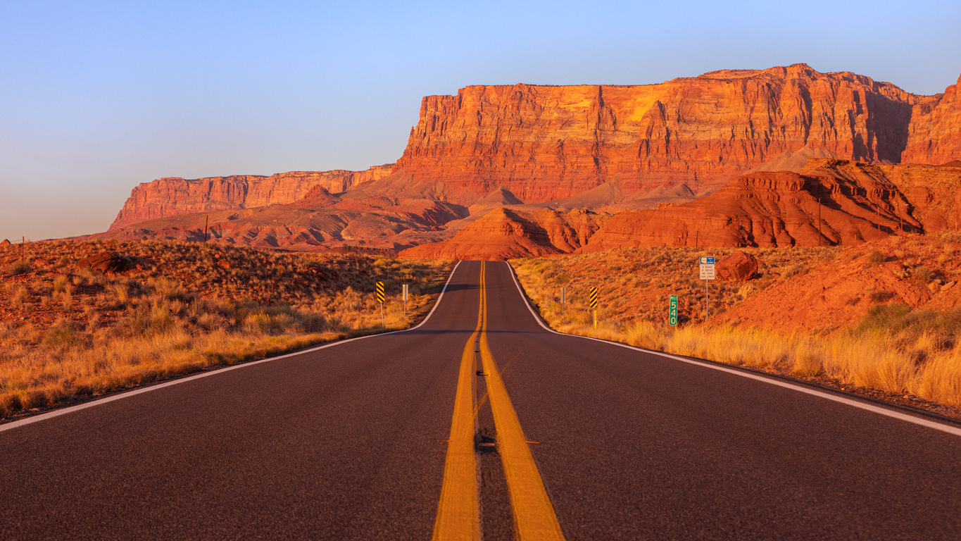 Eine gerade Straße führt durch rote Wüstenlandschaft in Richtung Grand Canyon.
