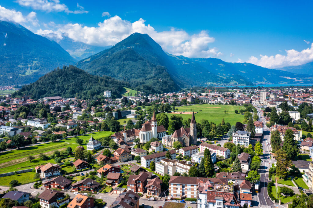 Luftaufnahme von Interlaken mit Kirchen, Wohnhäusern, grünen Wiesen und Alpenpanorama im Hintergrund.