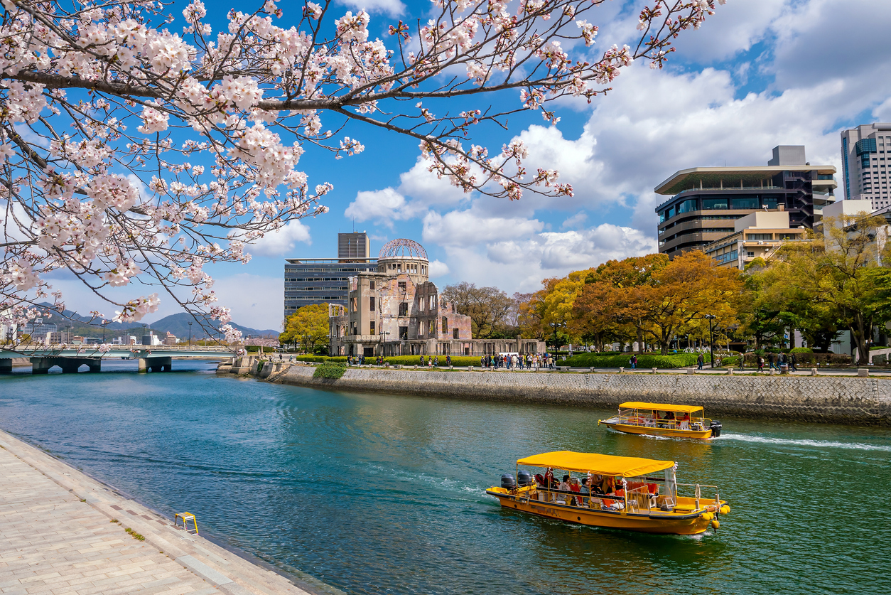 Kirschblüten rahmen den Genbaku Dome am Fluss in Hiroshima ein, während gelbe Boote vorbeifahren.