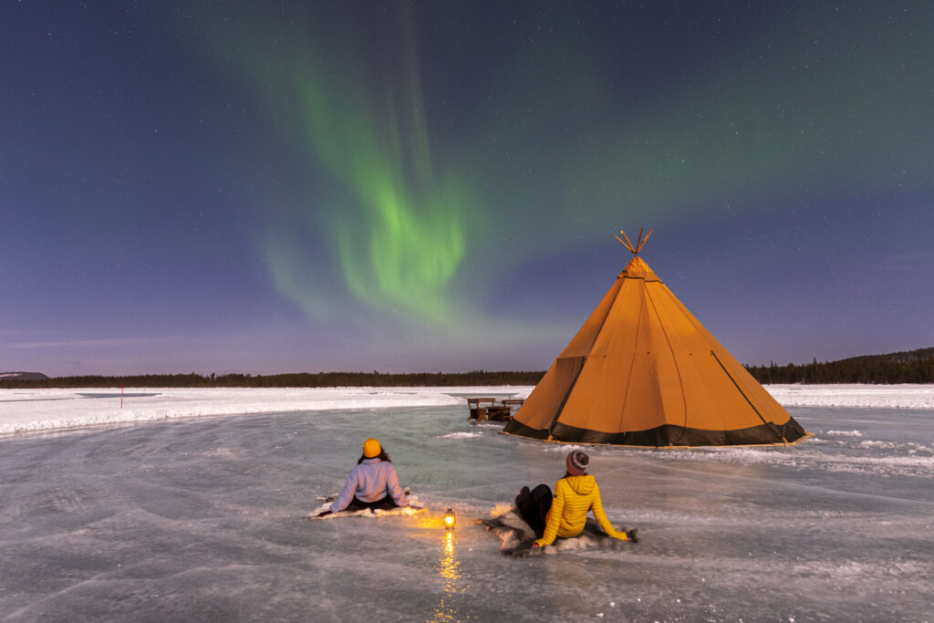 Zwei Personen sitzen auf einem gefrorenen See neben einem Zelt, während schwaches Polarlicht am Himmel schimmert.