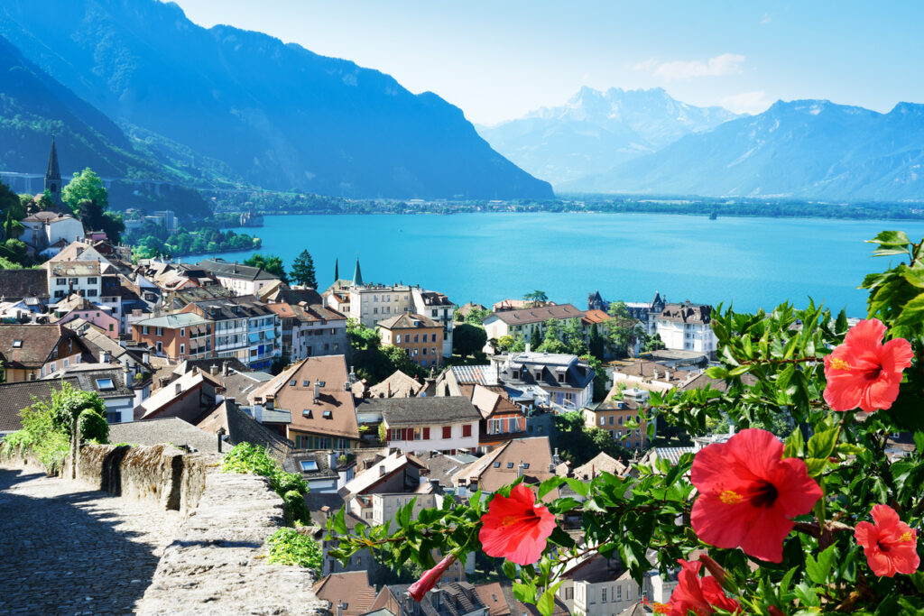 Eine Aussicht auf Montreux mit roten Blüten im Vordergrund, dem blauen See und Bergen im Hintergrund.