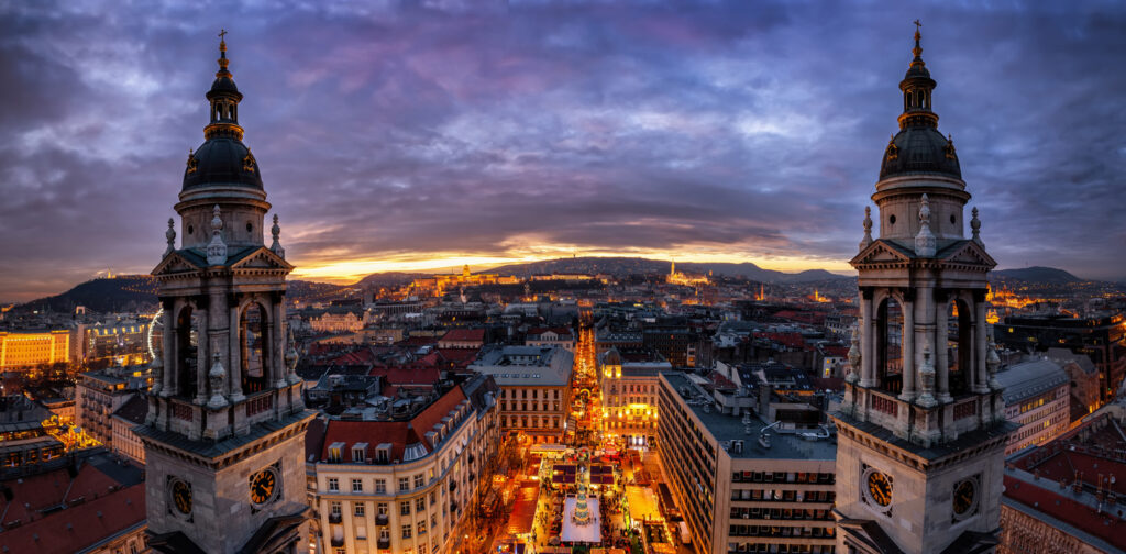 Von hoch oben sieht man Budapest mit zwei Kirchtürmen und einer langen, hellen Straße bis zum Horizont.