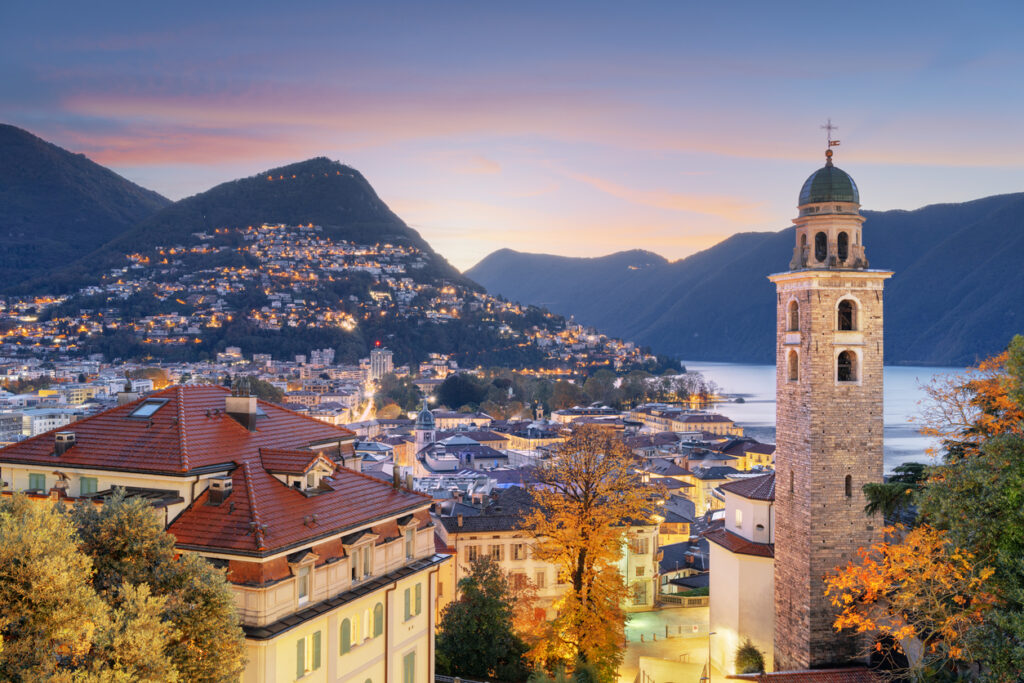 Blick über Lugano in der Dämmerung mit beleuchtetem Kirchturm, Stadtlichtern, See und Bergen.