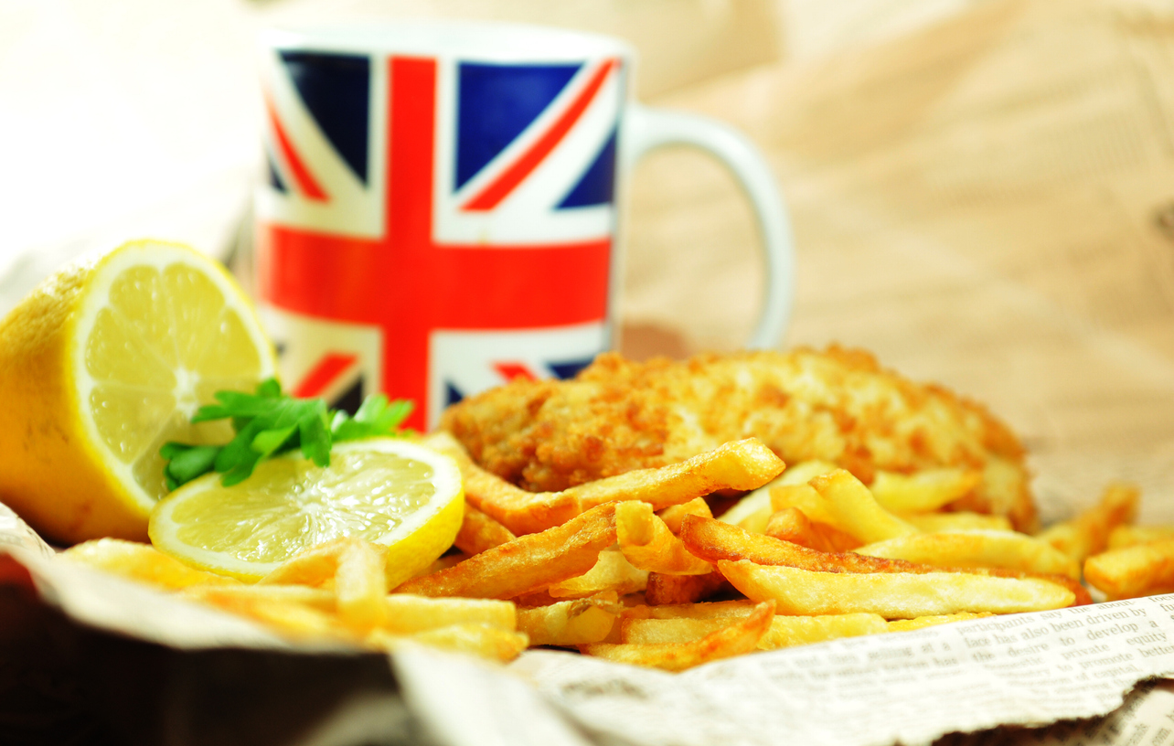 Fish and Chips mit Zitronenscheiben liegt in Papier, daneben steht eine Tasse mit Union-Jack-Motiv.