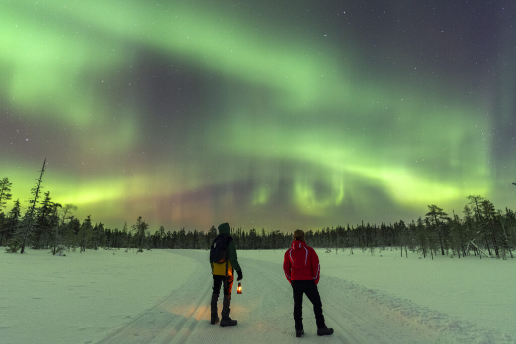 Zwei Menschen stehen auf einer verschneiten Strecke in Lappland und beobachten grüne Polarlichter am Sternenhimmel.