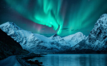 Kräftige Polarlichtbänder stehen über schneebedeckten Bergen und einem dunklen See.