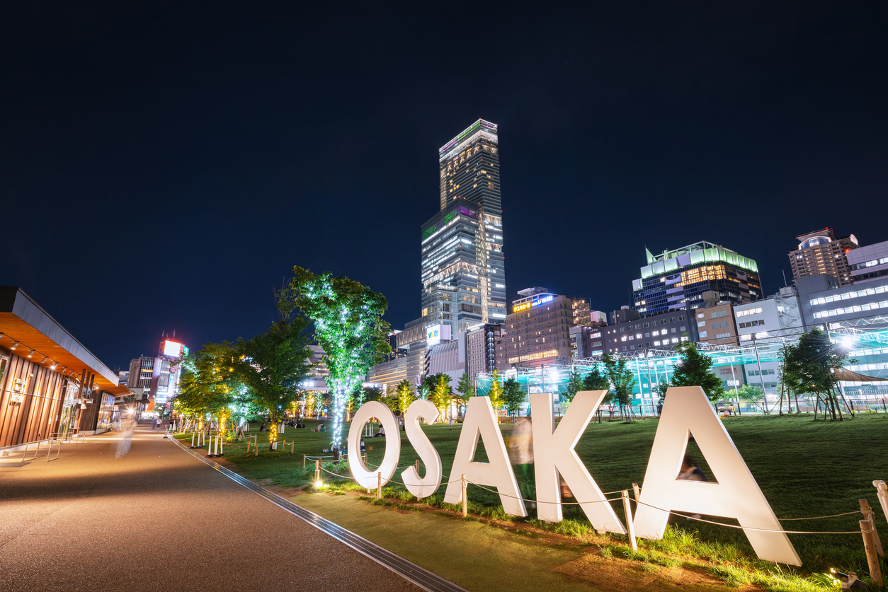 Ein großer OSAKA-Schriftzug steht nachts vor der beleuchteten Skyline von Osaka.