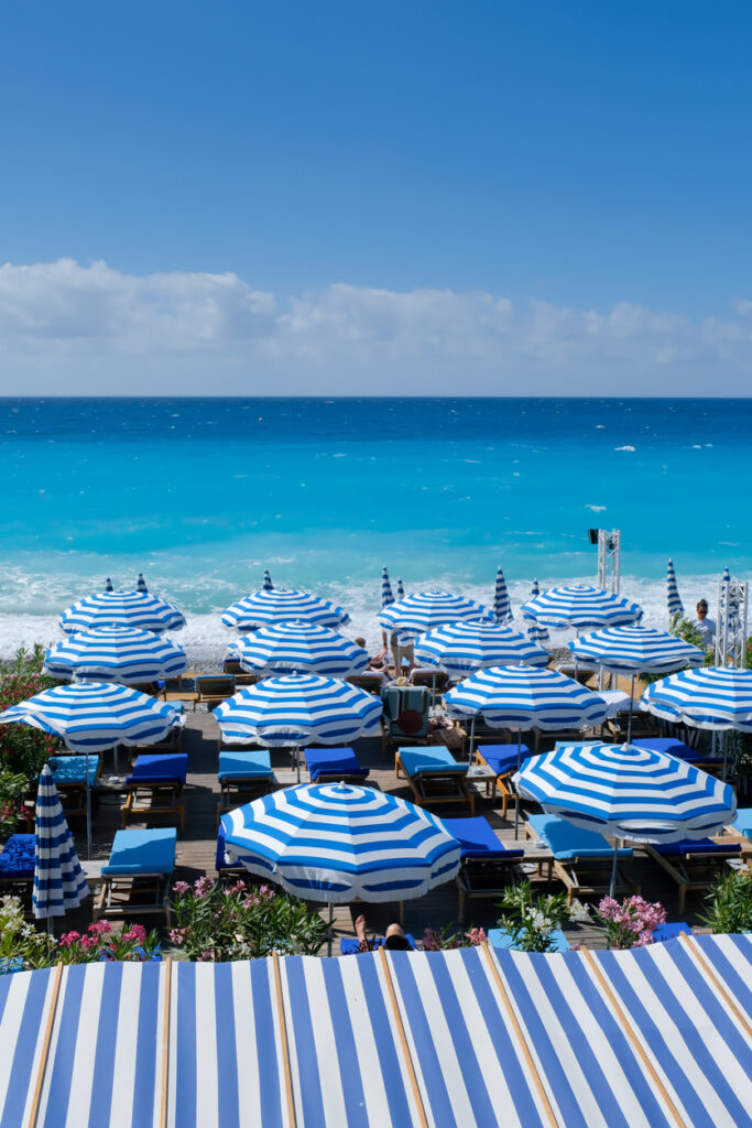 Viele Sonnenschirme stehen vor dem Meer mit kräftigem Blau und weißen Wellen.
