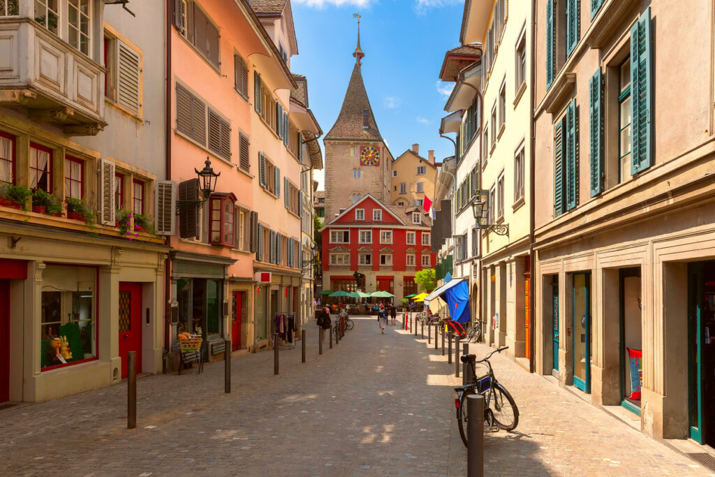 Schmale Gasse in Zürich mit historischen Häusern, Fahrrädern und einem Turm im Hintergrund.