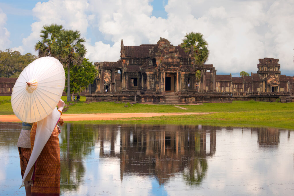 Eine Person mit hellem Schirm steht am Wasser, dahinter erhebt sich eine historische Tempelanlage.