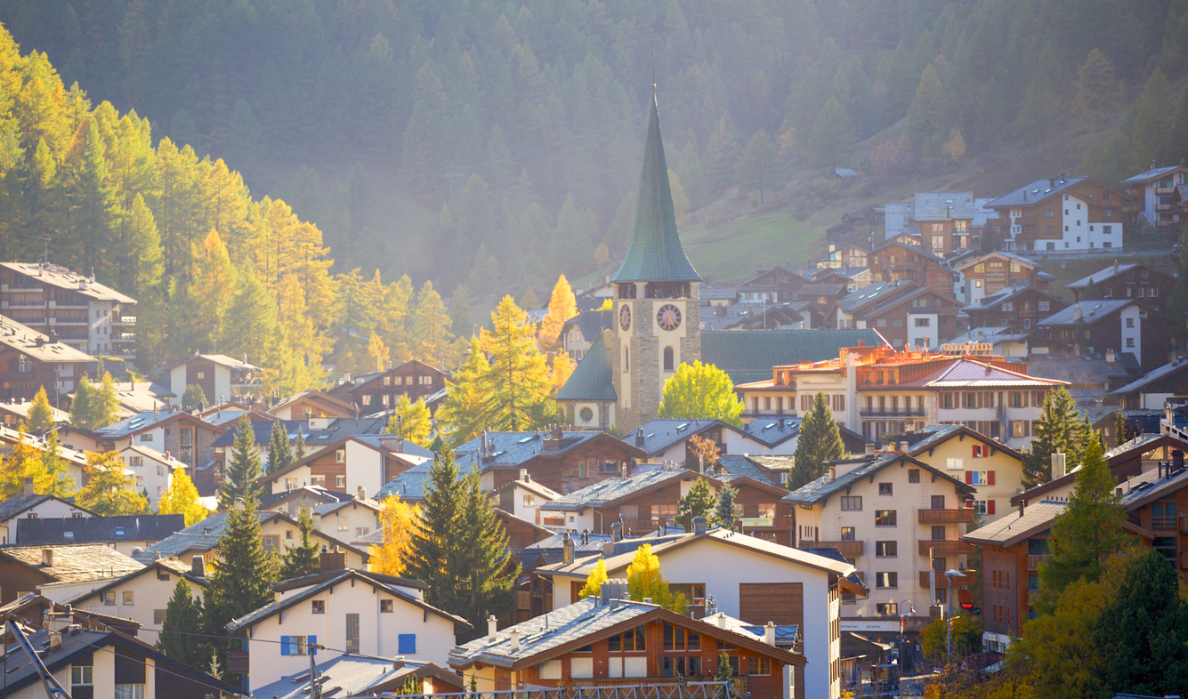Blick über Zermatt mit Kirche und Chalets vor herbstlich gelb leuchtenden Bäumen am Berghang.