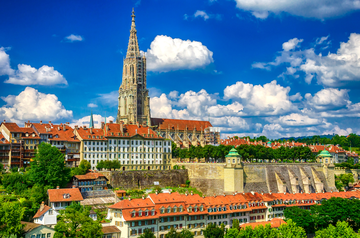 Das Berner Münster ragt über roten Dächern und der Aare unter einem wolkigen Himmel auf.
