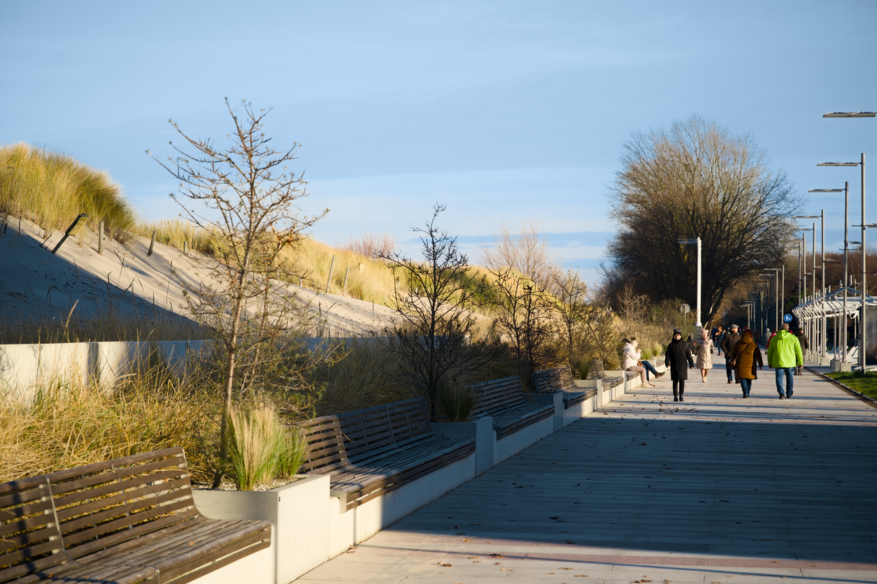 Spaziergänger laufen auf der breiten Promenade neben Dünen, Bänken und Laternen in Kolberg.