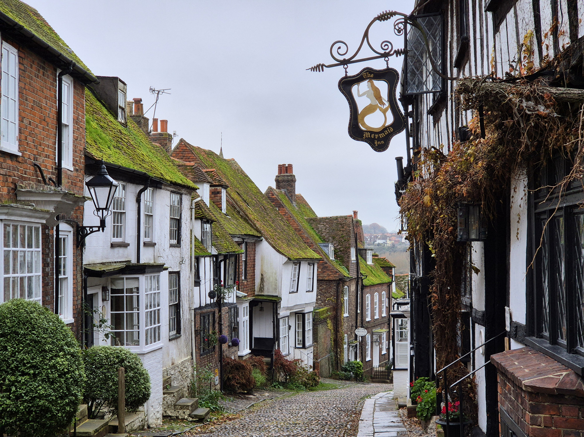 Enge Kopfsteinpflasterstraße mit alten Häusern und moosigen Dächern in einem englischen Dorf.
