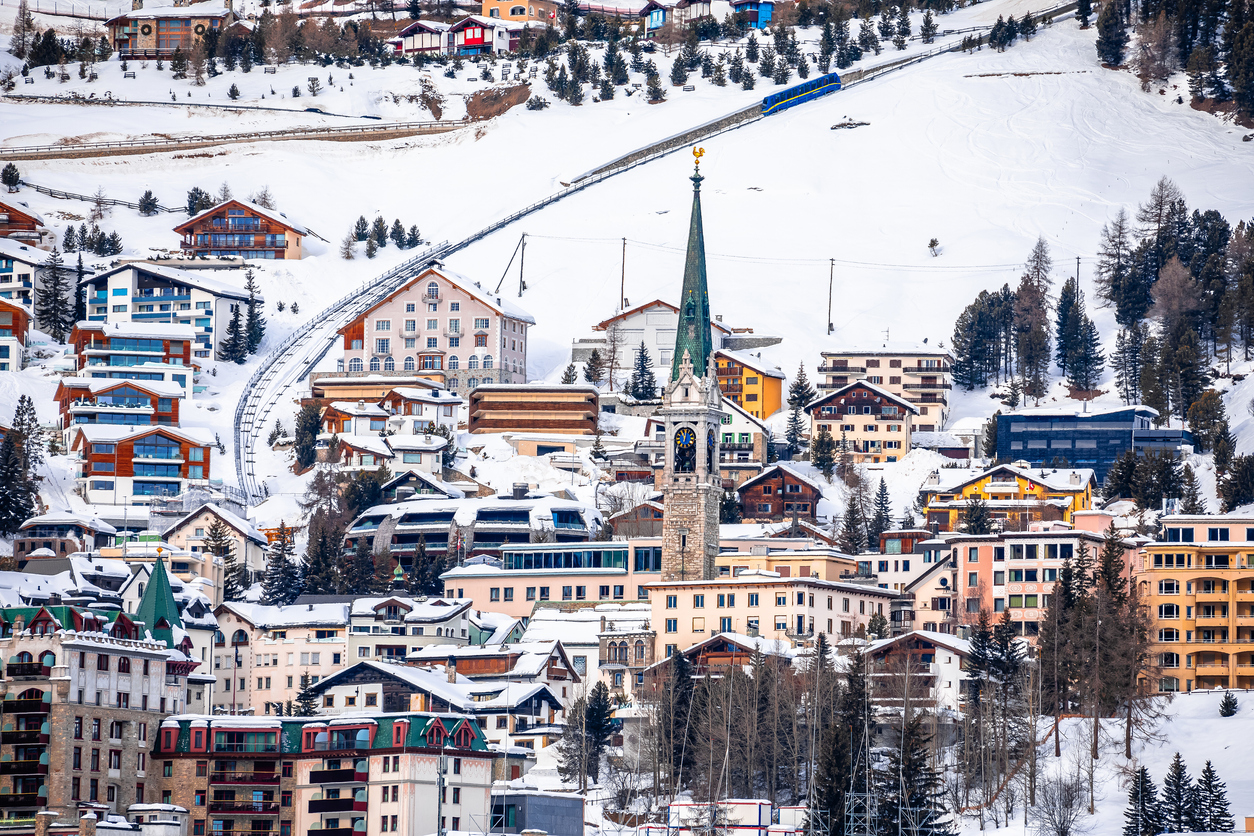 Verschneites St. Moritz mit markantem Kirchturm, Chalets und einem blauen Zug, der oberhalb des Ortes fährt.