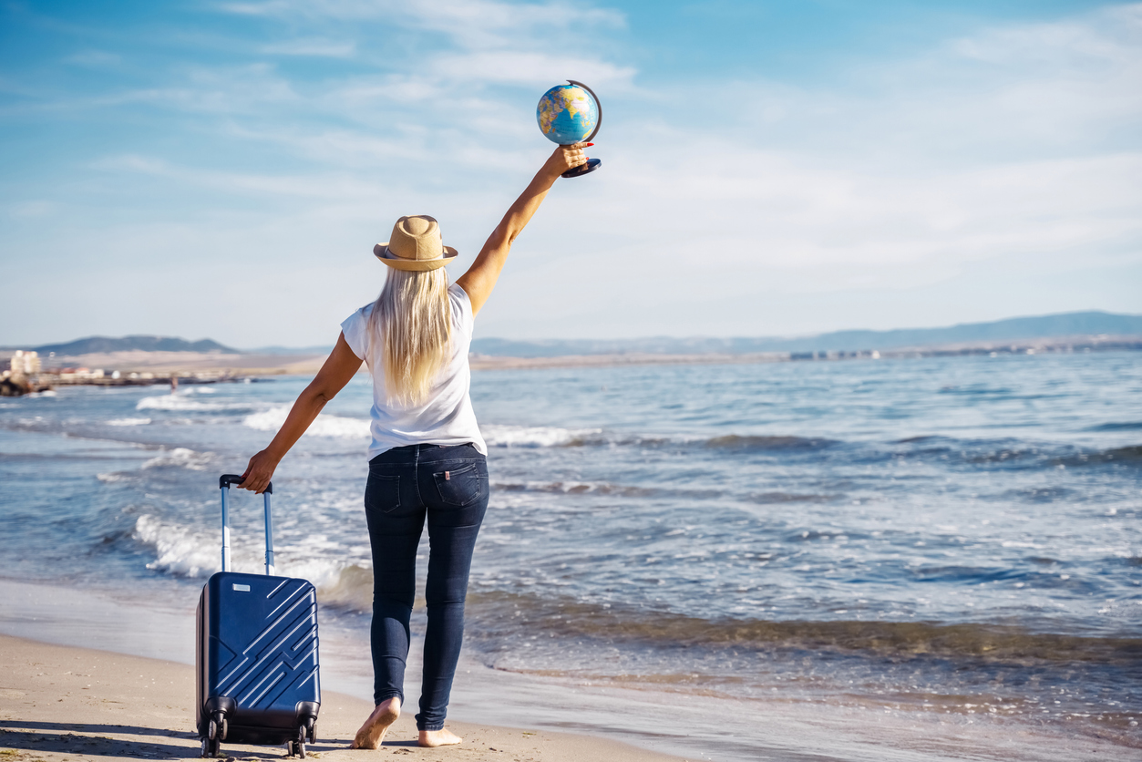 Eine Frau mit Koffer hält am Strand einen Globus hoch als Symbol für Urlaub in Bulgarien.