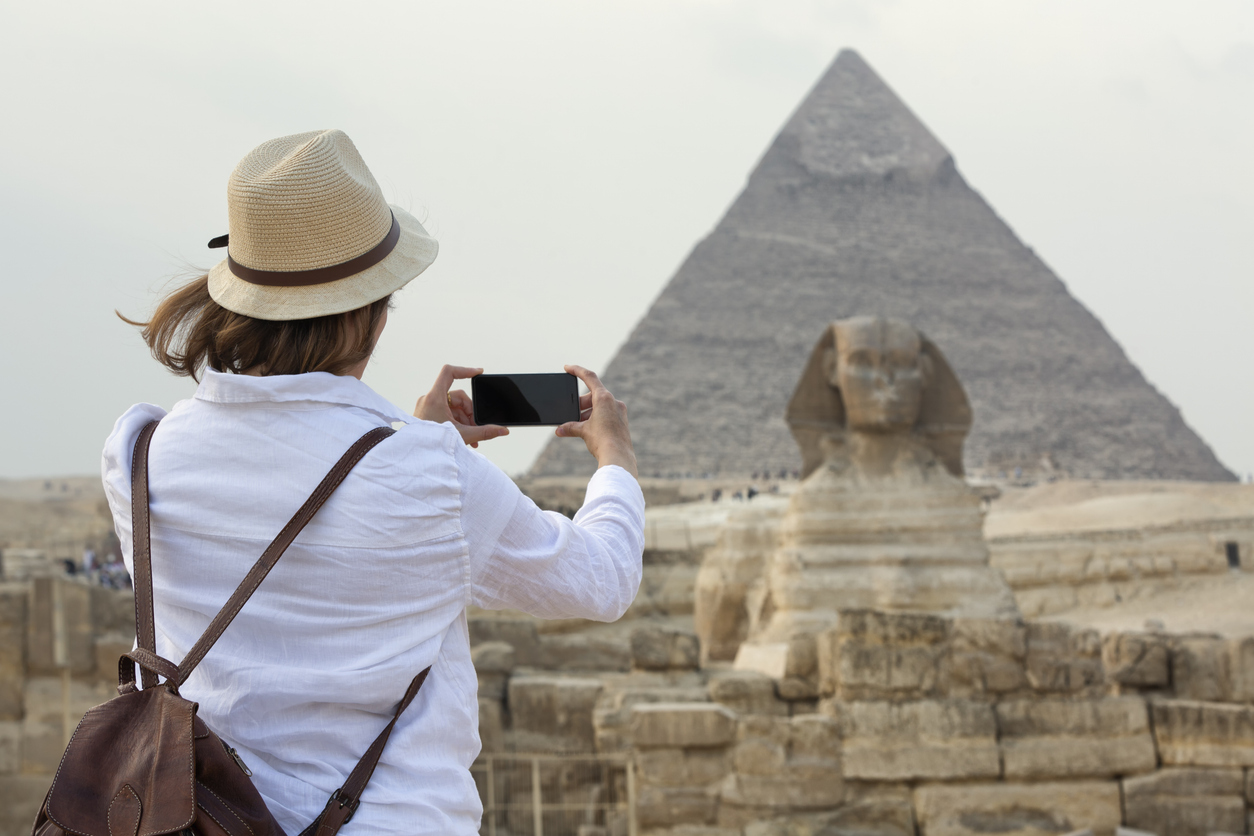 Eine Touristin hält ein Smartphone hoch und fotografiert die Sphinx mit einer Pyramide im Hintergrund.