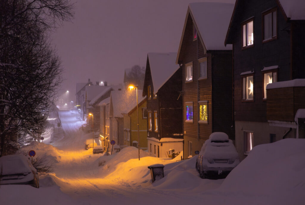 Verschneite Straße in Tromsø bei Nacht mit beleuchteten Häusern und Straßenlampen.