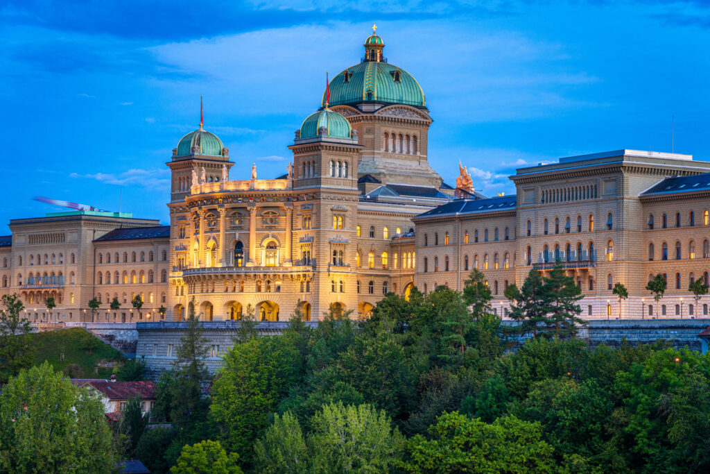 Das Bundeshaus mit grünen Kuppeln leuchtet vor blauem Himmel über einer Baumreihe.