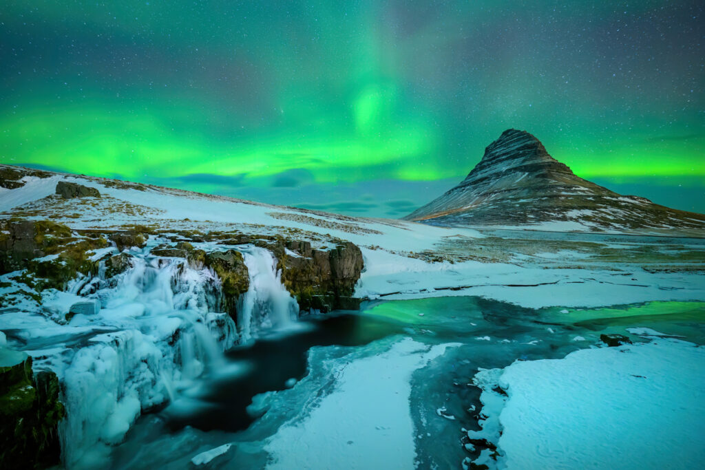 Grünes Polarlicht leuchtet über einem vereisten Wasserfall und einem markanten Berg.