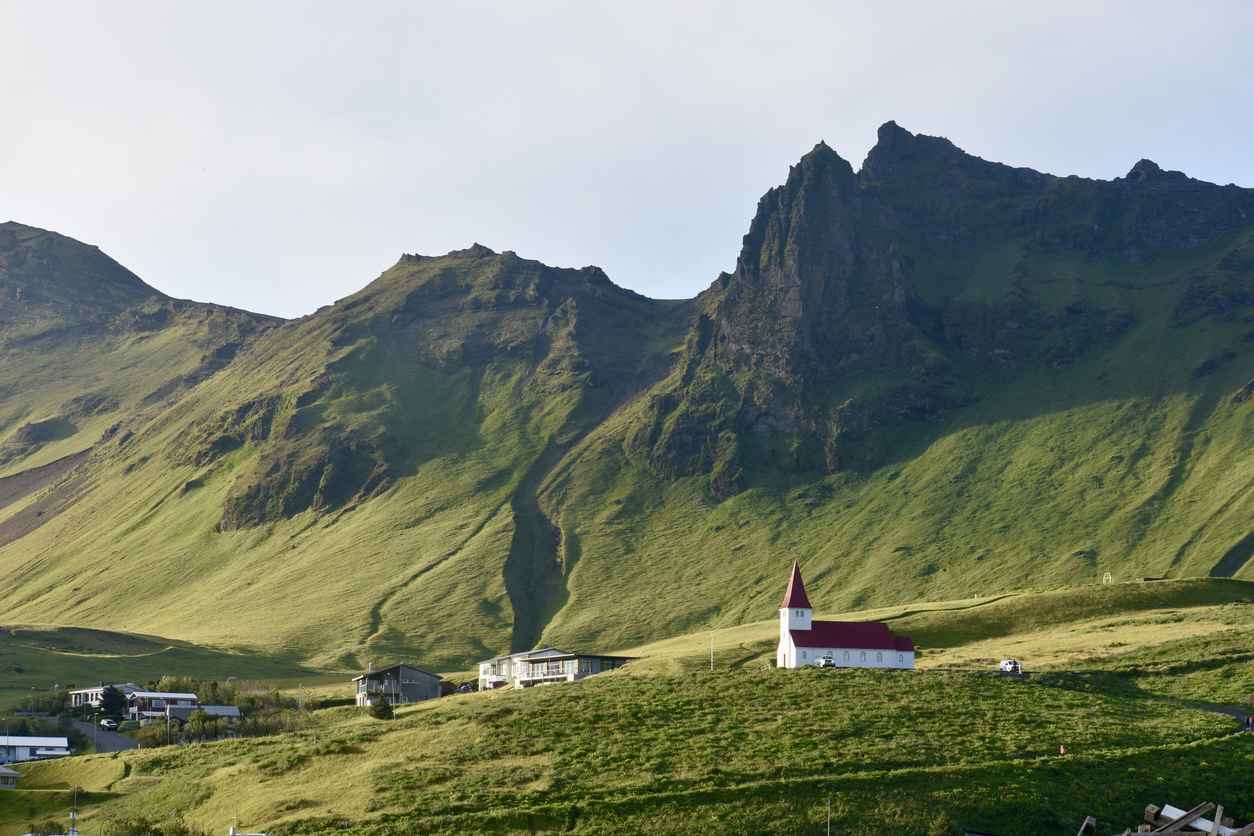 Die Kirche von Vík í Mýrdal steht mit rotem Dach vor steilen, grünen Berghängen.