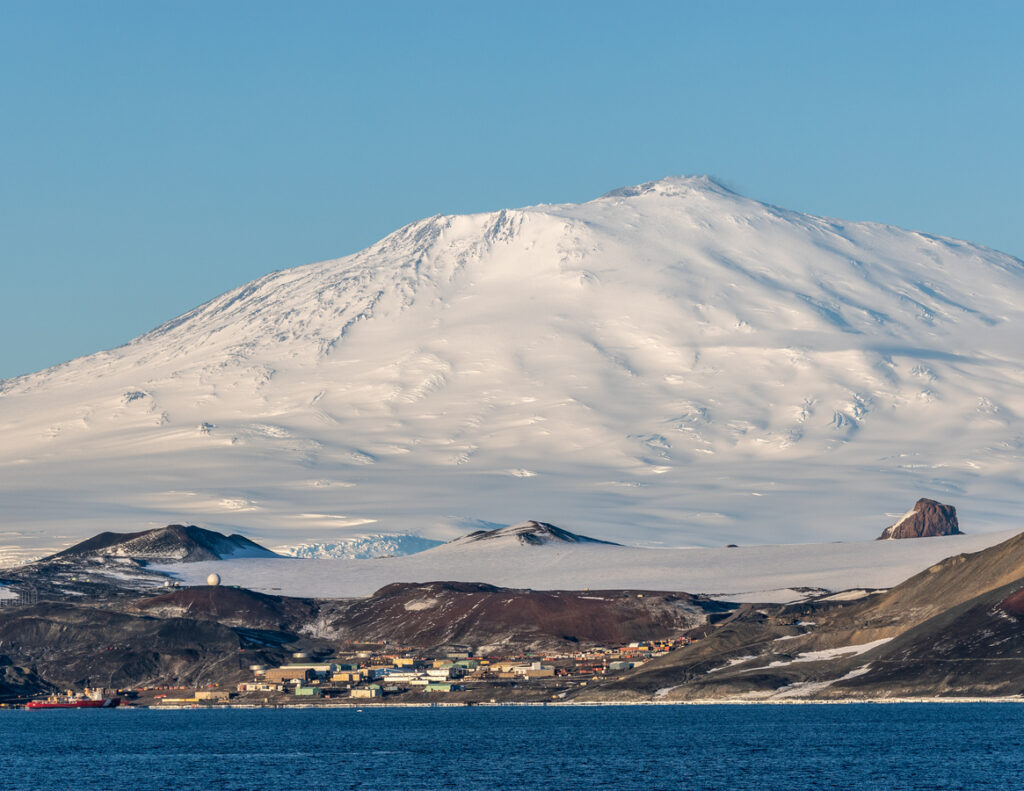 McMurdo Station liegt am Meer, dahinter erhebt sich ein riesiger schneebedeckter Berg unter blauem Himmel.