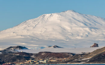McMurdo Station liegt am Meer, dahinter erhebt sich ein riesiger schneebedeckter Berg unter blauem Himmel.