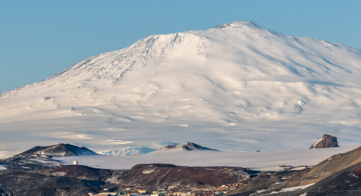 McMurdo Station liegt am Meer, dahinter erhebt sich ein riesiger schneebedeckter Berg unter blauem Himmel.