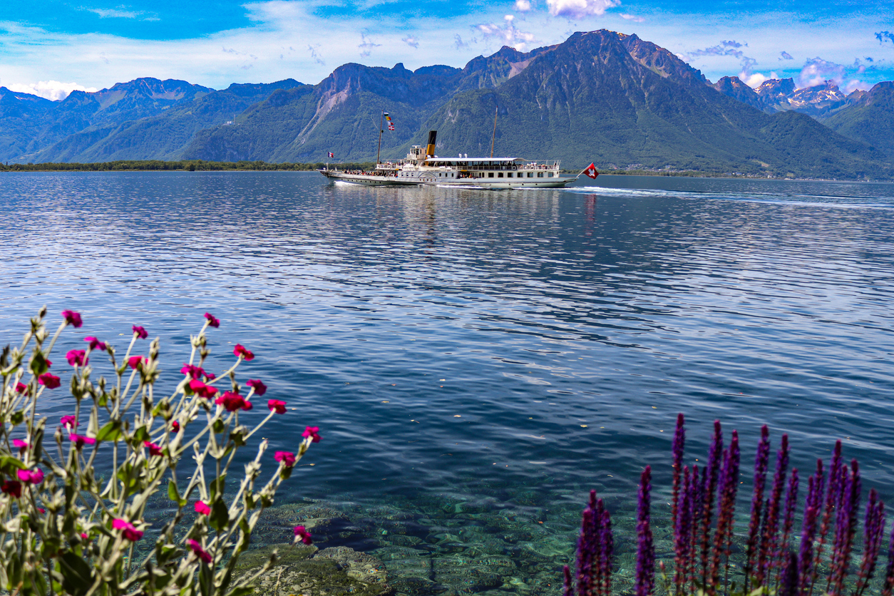 Ein historisches Dampfschiff fährt über den Genfersee, während im Hintergrund die Berge und am Ufer Blumen zu sehen sind.