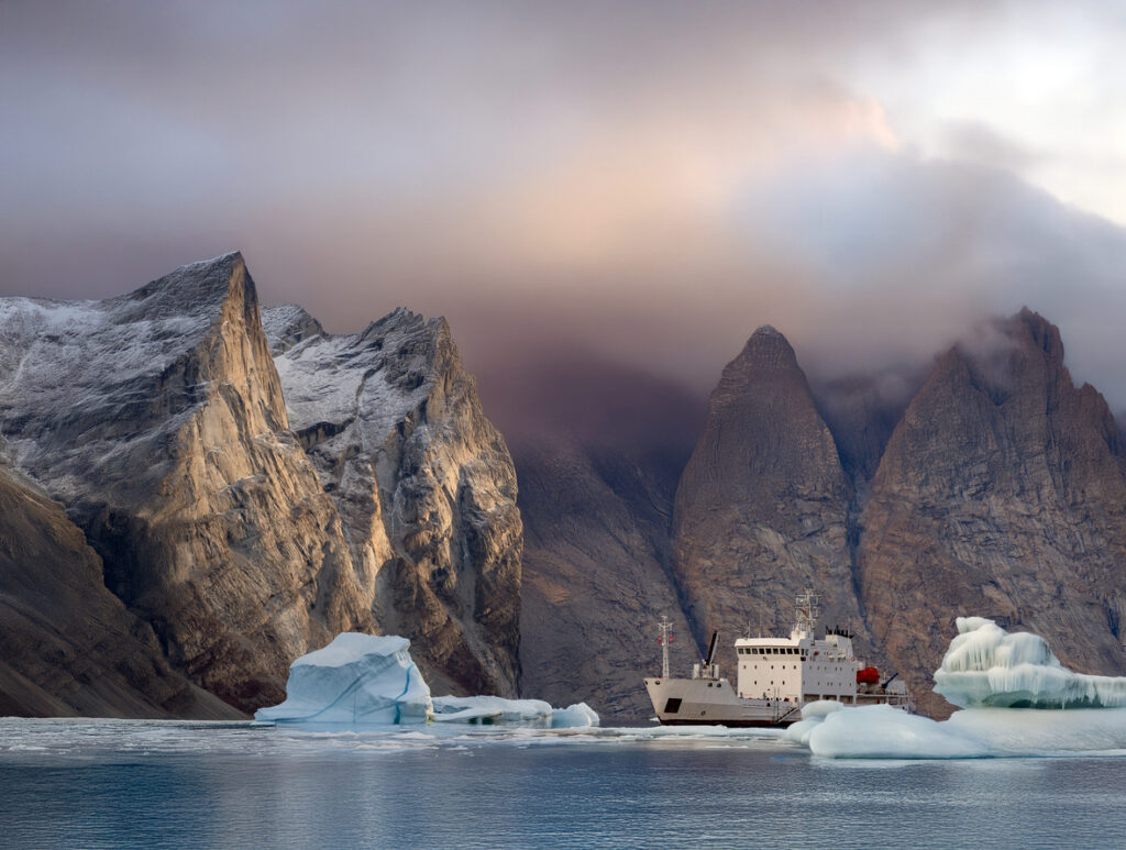 Expeditionsschiff vor hohen Bergen und treibendem Eis auf ruhigem Wasser.