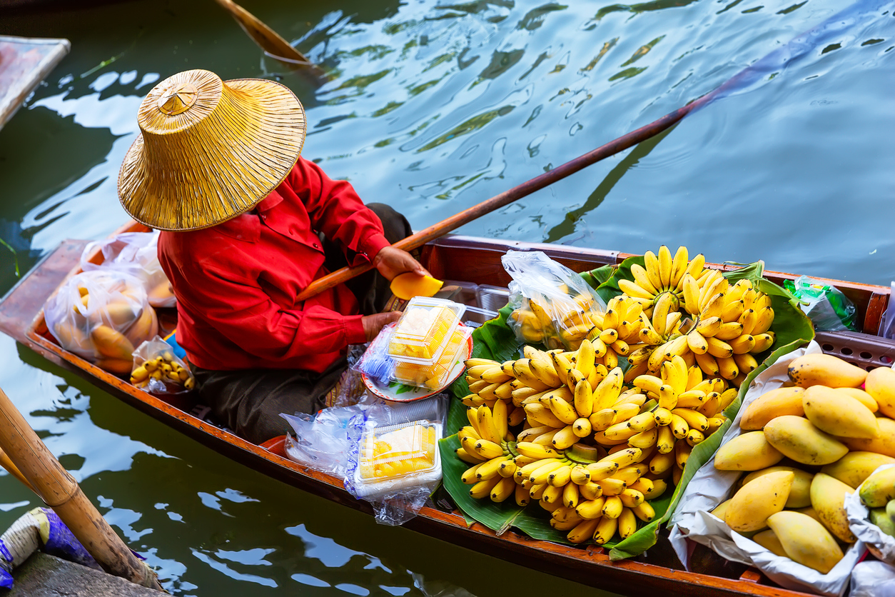 Eine Person mit Strohhut sitzt in einem Holzboot neben großen Bündeln Bananen und Mangos.