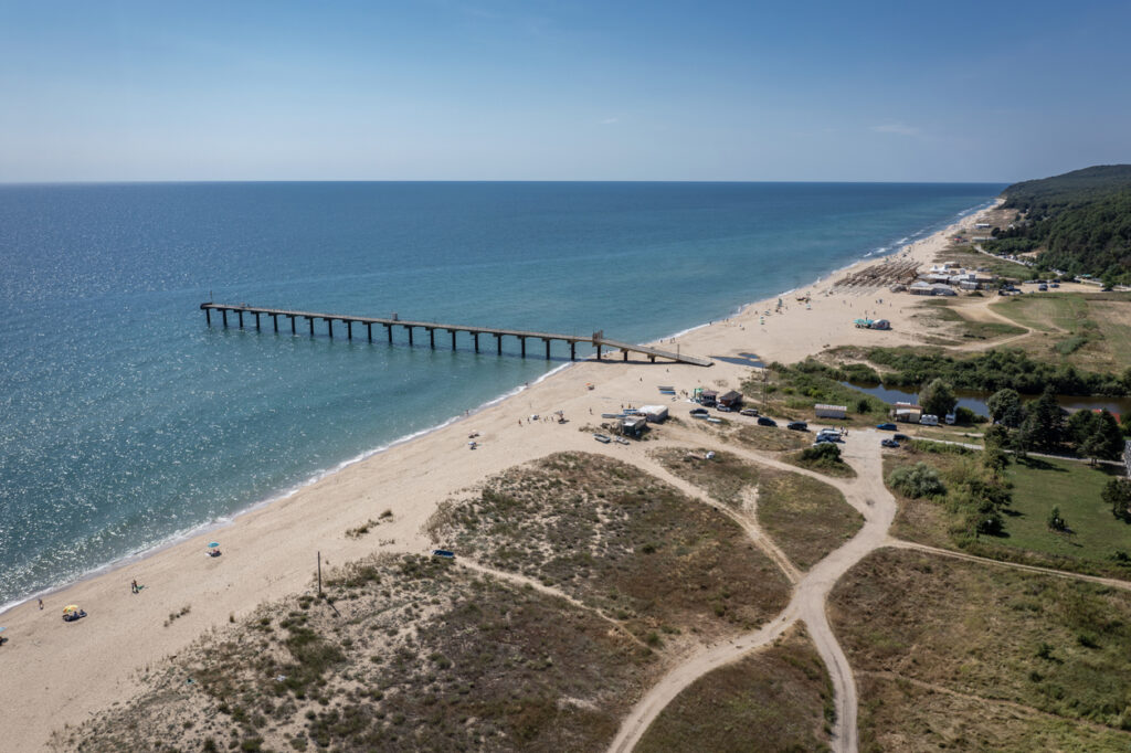 Luftaufnahme vom Strand von Shkorpilovtsi mit langem Sandstrand, blauem Meer, einem Steg und wenigen Strandgästen am Ufer.