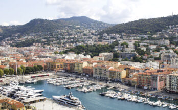 Ein Hafenbecken liegt vor einer Reihe farbiger Häuser und grüner Hügel.