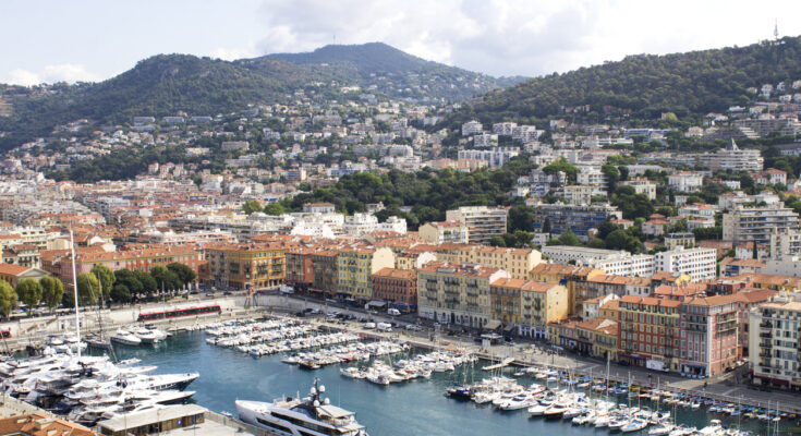 Ein Hafenbecken liegt vor einer Reihe farbiger Häuser und grüner Hügel.