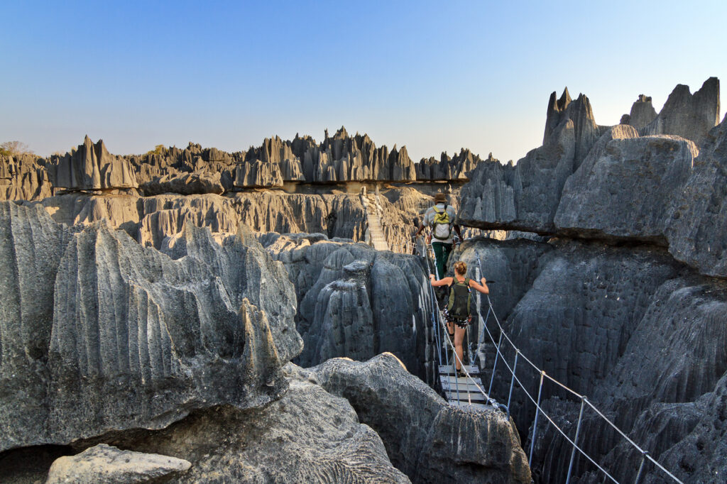 Zwei Wanderer überqueren eine schmale Hängebrücke in einer Landschaft aus spitzen grauen Felsnadeln.
