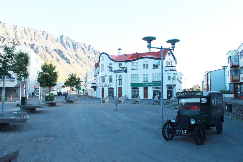 Eine kleine Straße in Ísafjörður mit weißen Häusern, Bergkulisse und einem alten Lieferwagen am Rand.