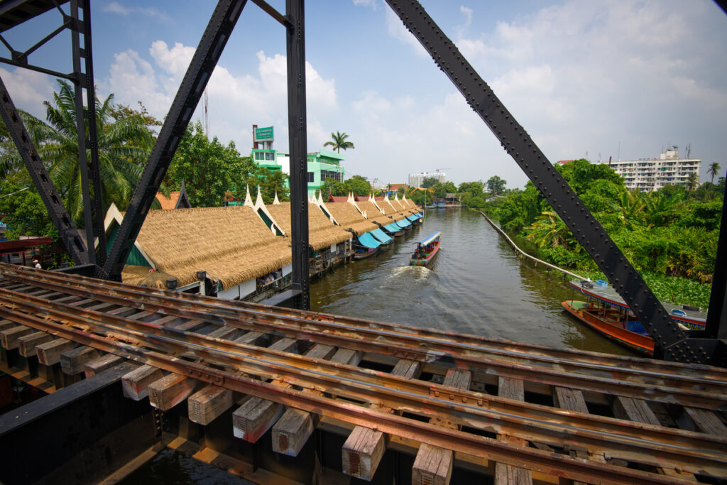 Boote fahren unter einer Stahlbrücke entlang des Kanals beim Taling Chan Floating Market in Bangkok.