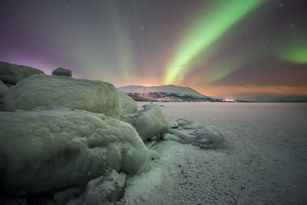 Grüne Polarlichter ziehen über eine eisige Uferlandschaft mit Blick auf die Berge bei Abisko.