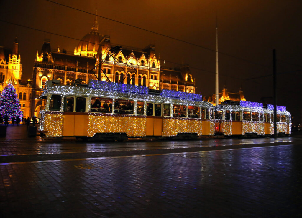 Eine beleuchtete Straßenbahn fährt am Ufer entlang, während das Parlament in Budapest golden strahlt.