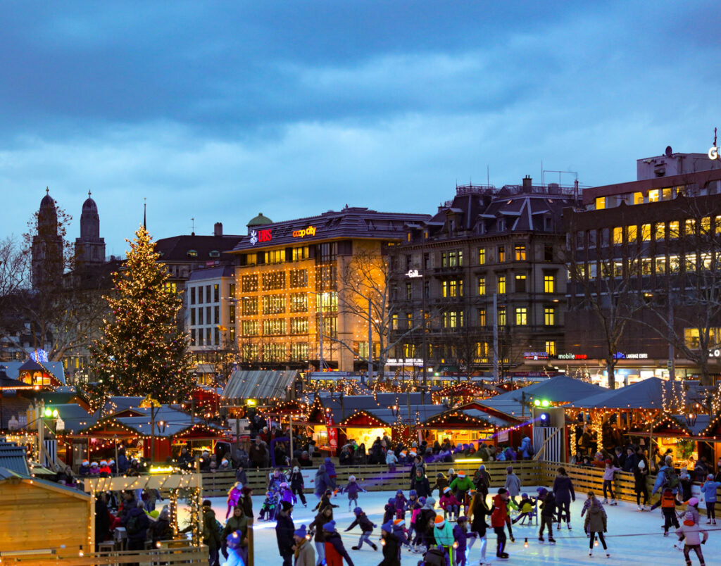 Weihnachtsmarkt in Zürich mit beleuchteten Buden, großem Baum und vielen Menschen auf einer Eislaufbahn.