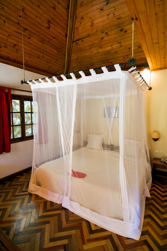 Ein Bett mit weißem Moskitonetz hängt in einem warm eingerichteten Zimmer mit Holzdecke.