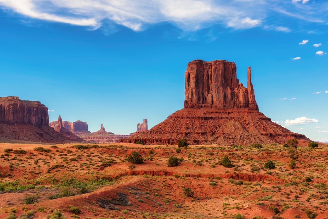 Ein gewaltiger Sandstein-Monolith ragt in Monument Valley aus der roten Ebene unter blauem Himmel auf.