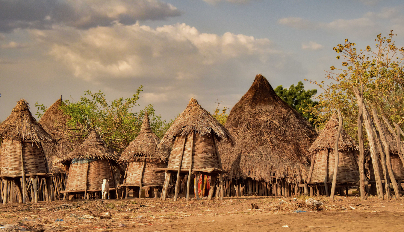 Mehrere traditionelle Rundhütten mit Strohdächern stehen auf trockenem Boden vor einem bewölkten Himmel.
