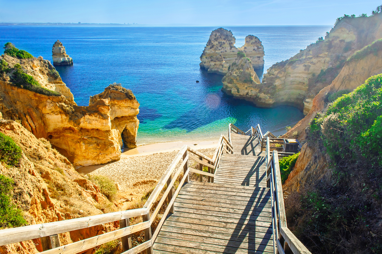 Eine Holztreppe führt zwischen goldenen Felsen zu einer kleinen Sandbucht mit türkisblauem Meer an der Algarve.