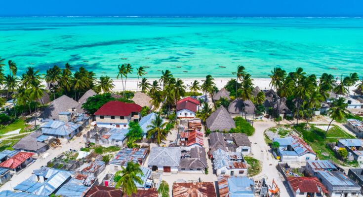 Ein Dorf mit Dächern aus Blech und Palmen liegt direkt an einer türkisfarbenen Küste auf Sansibar.