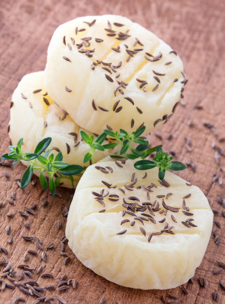 Kleine runde Käsestücke liegen mit Kümmel und einem Thymianzweig auf einem Holzbrett.