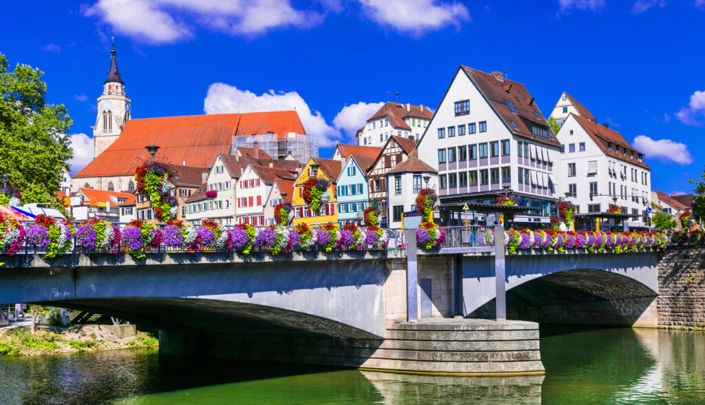 Eine Brücke mit üppigem Blumenschmuck überspannt den Fluss vor der Altstadtkulisse mit Kirchturm.